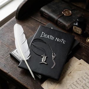 Può includere: Un taccuino nero con la scritta Death Note stampata in bianco, dotato di un ciondolo argentato a forma di lettera L su un cordino nero e una penna a forma di piuma bianca. Il set è disposto con una boccetta etichettata Kira's Ink.