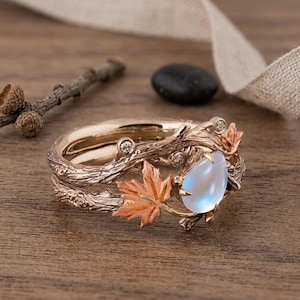 Peut inclure: Une bague en or rose avec un anneau texturé imitant des branches d'arbre. Elle est ornée de petites feuilles d'érable métalliques et d'une pierre de lune centrale en forme de poire, accentuée par de minuscules pierres rondes serties.
