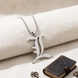 May include: A silver-toned metal pendant shaped like the letter L in a stylised, gothic-inspired font, suspended from a matching silver-toned ball chain necklace. The pendant has a smooth, reflective surface and a distinct, sharp-edged design.