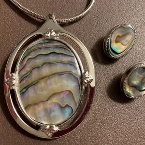 May include: A silver-toned jewelry set featuring an oval pendant and matching earrings. The pendant and earrings showcase iridescent abalone shells, with a range of colors including blue, green, and gold. The necklace has a silver chain.