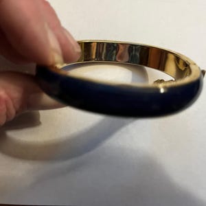 May include: A gold-toned bangle bracelet with a navy blue enamel band. The bracelet has a hinged clasp and a polished finish. This piece of jewelry is a classic accessory.