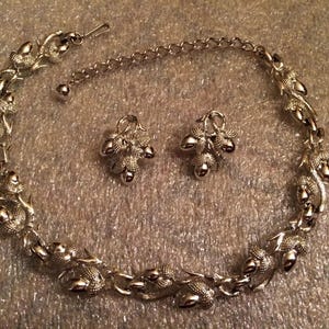 May include: Silver-toned necklace and matching earrings set. The necklace features a chain with decorative acorn-like elements. The earrings have a similar design. The jewelry set is displayed on a textured surface.
