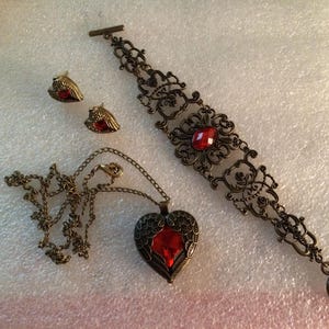 May include: A set of antique-style jewelry featuring a bronze-colored bracelet, necklace, and earrings. The bracelet and necklace pendant have a heart shape with a red gemstone. The earrings are small and have a similar design.
