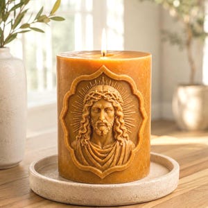 May include: A cylindrical beeswax candle in a warm golden-brown hue, featuring a detailed embossed relief of a figure with long hair and a crown of thorns centered within a decorative frame. The candle is lit, with a small flame at the top.