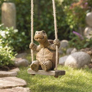 May include: A brown turtle figurine with a textured shell and skin, sitting on a small rectangular wooden swing suspended by two thick jute ropes. The turtle has its front limbs wrapped around the ropes, appearing to hold onto the swing.
