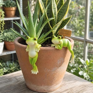 Può includere: Due statuette di rane verde chiaro appollaiate sul bordo di un vaso di terracotta contenente un'aloe vera e una sansevieria. Una rana pende verticalmente mentre l'altra è sdraiata accanto a un cartello in legno con scritto Save Water Upon Plants.