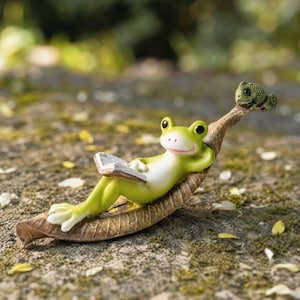 Könnte beinhalten: Eine hellgrüne Froschfigur, die auf einer braunen, geschwungenen, blattförmigen Basis liegt und ein offenes Buch hält. Eine kleine, texturierte grüne Chamäleonfigur sitzt auf der Spitze des Blattes und blickt in Richtung des Frosches.