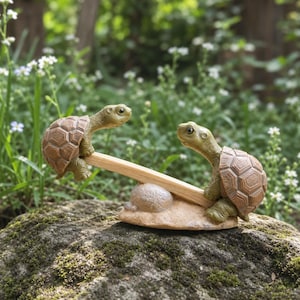 Op de afbeelding: Een decoratief tuinbeeldje met twee groene schildpadden met bruine schilden die op een wipwap balanceren. De wipwap wordt ondersteund door een centrale, steenachtige basis. De schildpadden zitten aan de uiteinden van de houten plank.
