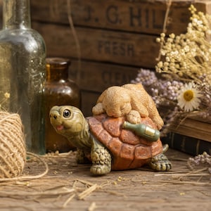 May include: A decorative figurine featuring a light brown rabbit sleeping on the back of a green and brown turtle. The turtle has a small green bottle resting on its shell. The figurine is crafted with detailed textures on the animal fur and shell.