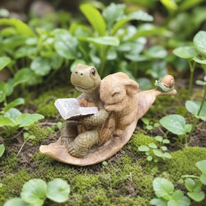 May include: A decorative figurine featuring a light green turtle holding an open book, with a small tan rabbit leaning against it and a tiny green snail perched on the end of a large, curved tan leaf base that supports all three figures.
