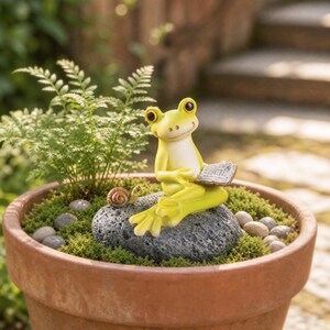 Puede incluir: Una figura de rana de color verde claro sentada sobre una roca gris oscuro texturizada, sosteniendo un pequeño libro abierto. Un diminuto caracol marrón está colocado junto a la rana sobre la roca. La figura está en una maceta de terracota con musgo.