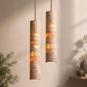 Peut inclure: Deux suspensions cylindriques dotées d'une finition en pierre beige poreuse et texturée. Chaque lampe est suspendue par un cordon fin et droit, diffusant une lueur chaude et douce qui souligne les motifs naturels et stratifiés de la pierre.
