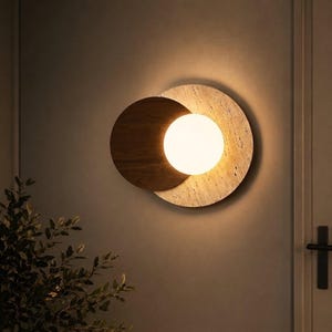 Op de afbeelding: Een wandlamp met een ronde beige stenen achterplaat met een natuurlijke textuur, gedeeltelijk overlapt door een kleinere donkerbruine ronde houten schijf, met een gloeiende witte bolvormige glazen lamp in het midden.