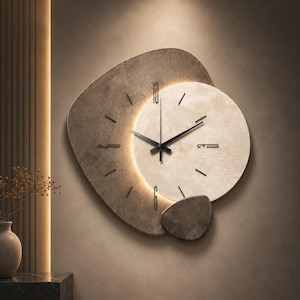 Peut inclure: Une horloge murale composée d'une base texturée gris foncé de forme irrégulière superposée à un cadran circulaire beige clair. Elle présente des aiguilles et des index noirs, avec un rétroéclairage LED chaud illuminant la section centrale.