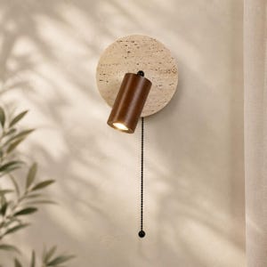 Peut inclure: Une applique murale composée d'une plaque arrière circulaire en pierre travertin beige et d'un spot cylindrique en bois brun foncé. La lampe est équipée d'une chaînette noire suspendue verticalement sous la tête du spot lumineux.
