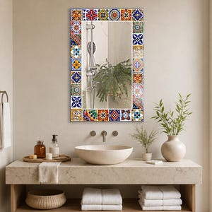 May include: A rectangular wall mirror featuring a decorative border composed of colorful square tiles with intricate geometric and floral patterns in shades of blue, yellow, red, green, and white, reflecting a shower head and a green plant.