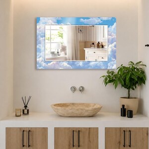 May include: A rectangular wall mirror featuring a decorative border printed with a blue sky and white clouds pattern. The mirror is mounted above a bathroom vanity, reflecting a window, white curtains, and a white cabinet with toiletries.