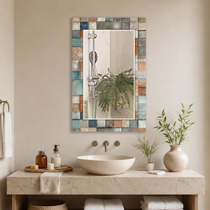 May include: A rectangular wall mirror featuring a decorative frame composed of small, square tiles in various shades of blue, copper, and grey. The mirror surface reflects a shower fixture and a green plant, set against a neutral wall.