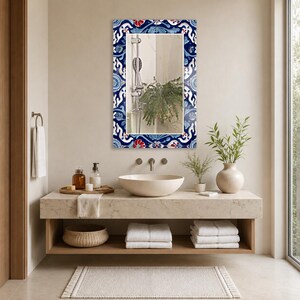 May include: A rectangular wall mirror featuring a decorative frame with a vibrant blue, white, and red floral pattern. The mirror reflects a shower head and a green plant, and it is mounted on a wall above a stone vanity with a basin sink.