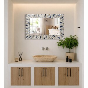 May include: A rectangular wall mirror featuring a decorative frame composed of small, multi-colored mosaic tiles in shades of blue, white, gray, and tan. The mirror reflects a bright room with a white vanity cabinet and a window with curtains.
