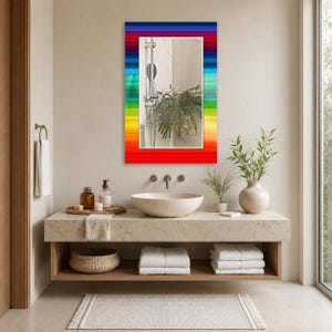 May include: A rectangular wall mirror featuring a vibrant, multi-colored border with horizontal stripes in shades of red, orange, yellow, green, blue, and purple. The reflective surface shows a shower head and a green plant in a bathroom setting.
