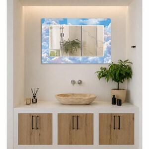 May include: A rectangular wall mirror featuring a decorative border printed with a blue sky and white clouds pattern. The reflective surface shows a bathroom interior with a shower head and a green plant, mounted above a stone vessel sink.