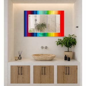May include: A rectangular wall mirror featuring a vibrant, multi-colored rainbow border with vertical stripes in shades of red, orange, yellow, green, blue, and purple. The mirror surface reflects a bathroom interior with a plant and fixtures.