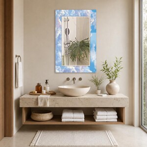 May include: A rectangular wall mirror featuring a decorative border printed with a blue sky and white clouds pattern. The reflective center shows a shower head and a green plant. The mirror is mounted on a wall above a bathroom vanity sink.
