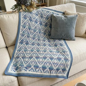 May include: A rectangular crochet blanket featuring a repeating geometric diamond pattern in shades of blue, teal, and white. The blanket has a solid blue border and is draped over a light-colored sofa next to a solid blue velvet pillow.