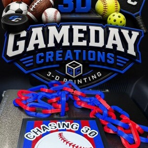 May include: A 3D printed sign featuring the text GAMEDAY CREATIONS and 3-D PRINTING, surrounded by various sports balls and a paddle. Below, a pentagonal plaque displays a baseball graphic with the text CHASING 30, accompanied by a blue and red plastic chain.