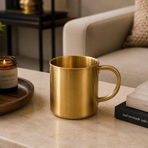 Könnte beinhalten: Ein zylindrischer Becher mit metallisch goldener Oberfläche und glattem, poliertem Finish. Der Becher verfügt über einen geschwungenen Henkel an der rechten Seite und eine weite Öffnung. Das Design ist schlicht und einheitlich mit messingartigem Glanz.