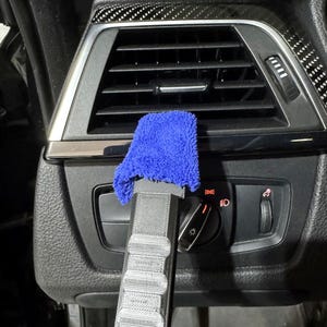 May include: A gray 3D-printed plastic cleaning tool with a blue microfiber cloth attachment, positioned against a black car air vent and headlight control panel featuring illuminated red icons and a circular dial for vehicle lighting settings.