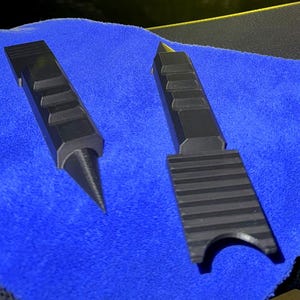 May include: Two black, rigid, elongated tools with textured grips. One tool features a sharp, pointed conical tip, while the other has a flat, wide end with a semi-circular cutout. Both items are matte black and have a geometric, industrial design.