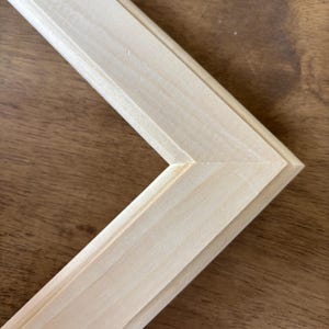 May include: A light-colored, unfinished wooden picture frame corner featuring a mitered joint. The frame has a smooth, pale wood grain texture with a stepped, tiered profile design that creates depth along the inner and outer edges of the frame.