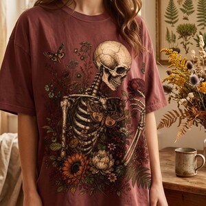 A person wearing an oversized mauve T-shirt printed with a gothic design of a skeleton surrounded by botanical flowers, sunflowers, and butterflies.