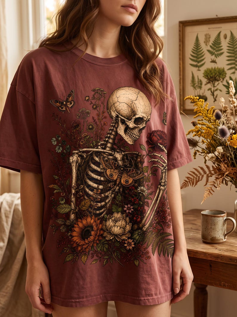 A person wearing an oversized mauve T-shirt printed with a gothic design of a skeleton surrounded by botanical flowers, sunflowers, and butterflies.