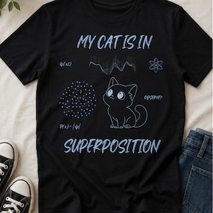 Pode incluir: Uma t-shirt preta de manga curta com um gráfico azul claro de um gato, fórmulas científicas e o texto "MY CAT IS IN SUPERPOSITION" juntamente com "OBSERVE?", "P(x)=/ψ/" e "ψ(x,t)" impressos na frente da peça.