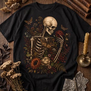 A black T-shirt laid flat on a wooden surface, featuring a graphic print of a skeleton amidst flowers and butterflies, accompanied by books, a candle, and dried flowers.