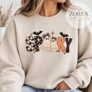 Pode incluir: Uma camisola bege de gola redonda com a palavra SPOOKY em estilo patchwork. As letras têm padrões de xadrez preto e branco, flores pretas e xadrez vichy laranja. Inclui dois fantasmas cobertos de renda com laços e morcegos pretos.