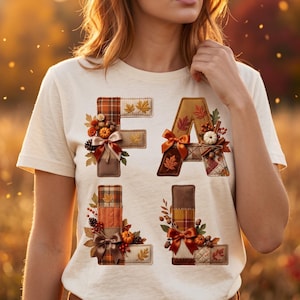 May include: A cream-coloured short-sleeve t-shirt featuring the word FALL spelled out in large, block-style letters. Each letter is decorated with autumn-themed elements including plaid fabric patterns, small pumpkins, pinecones, and satin bows.