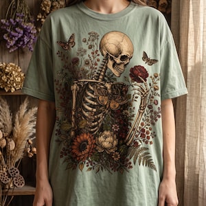 A person wearing a sage green oversized T-shirt displaying a vintage-style graphic of a skeleton interacting with botanical floral elements and butterflies.