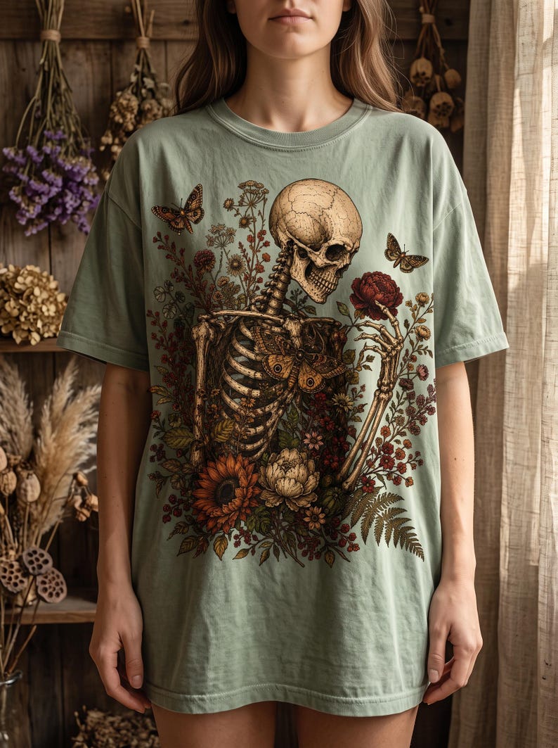 A person wearing a sage green oversized T-shirt displaying a vintage-style graphic of a skeleton interacting with botanical floral elements and butterflies.