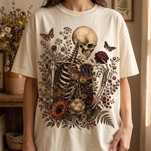 A person modeling a cream-colored T-shirt featuring a gothic skeleton graphic surrounded by dark floral and botanical artwork.