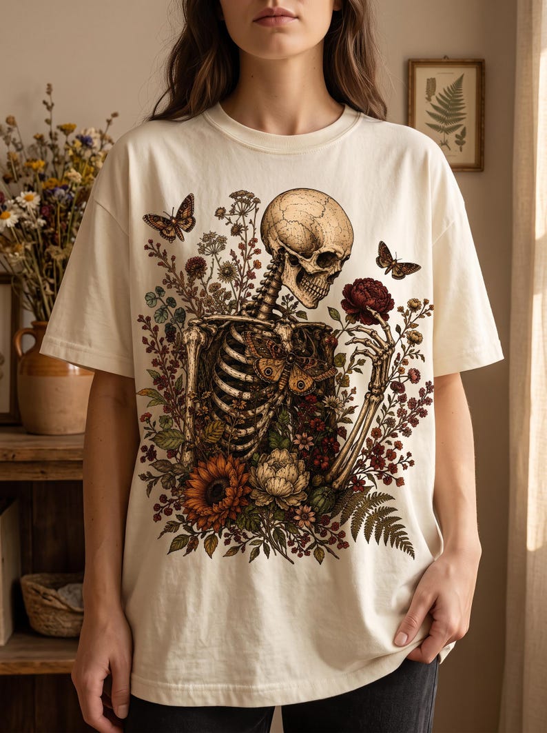 A person modeling a cream-colored T-shirt featuring a gothic skeleton graphic surrounded by dark floral and botanical artwork.