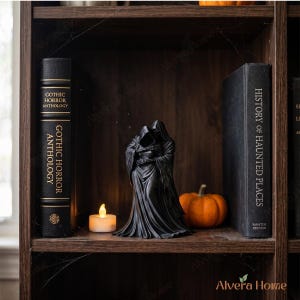 May include: A black figurine of two cloaked skeletal figures embracing, positioned between a book titled Gothic Horror Anthology and a book titled History of Haunted Places, with a small orange pumpkin and a white LED tea light candle nearby.