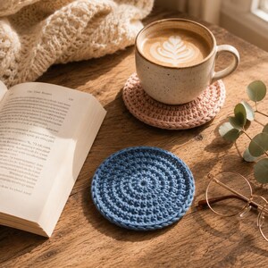 Könnte beinhalten: Zwei runde, handgehäkelte Untersetzer in den Farben Pastellrosa und Staubblau. Der rosa Untersetzer trägt eine Keramiktasse mit Kaffee und Latte Art, während der blaue Untersetzer flach neben einem aufgeschlagenen Buch auf der Oberfläche liegt.