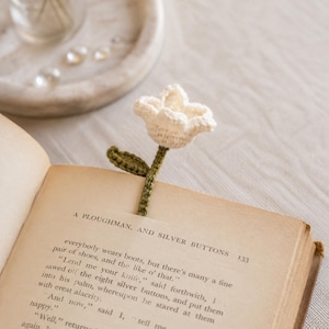 Könnte beinhalten: Ein cremefarbenes, gehäkeltes Blumen-Lesezeichen mit grünem Stiel und Blatt, das zwischen den Seiten eines offenen Buches steckt. Die Buchseite zeigt den gedruckten Text A PLOUGHMAN, AND SILVER BUTTONS und die Zahl 133 oben rechts.