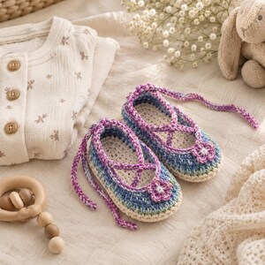 Könnte beinhalten: Ein Paar handgehäkelte Babyschuhe mit einer Basis aus meliertem blau-grünem Garn und cremefarbenen Sohlen. Die Schuhe haben violette geflochtene Schnürsenkel, die sich oben kreuzen, und sind mit einer kleinen violett-weißen Blumenapplikation verziert.