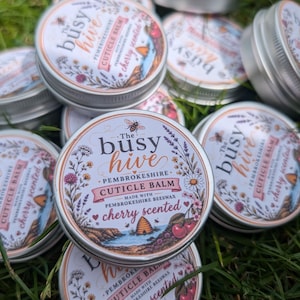 May include: Multiple round silver metal tins of The Busy Hive Pembrokeshire Cuticle Balm. Each lid features a white label with floral illustrations, a bee, a beehive, and the text: The Busy Hive Pembrokeshire Cuticle Balm cherry scented.