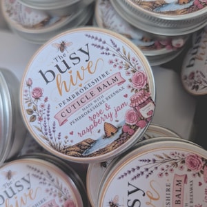 May include: A round metal tin of The Busy Hive Pembrokeshire Cuticle Balm with a white label featuring illustrations of a bee, lavender, roses, a beehive, and a jar of rose and raspberry jam, with text detailing the ingredients and brand name.